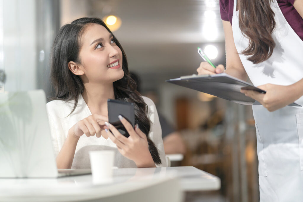 asian-female-customer-order-food-drink-from-smartphone-digital-menu-social-contactless-waitress-staff-smiling-millennial-waitress-with-paper-chart-taking-order-from-young-asian-female-customer